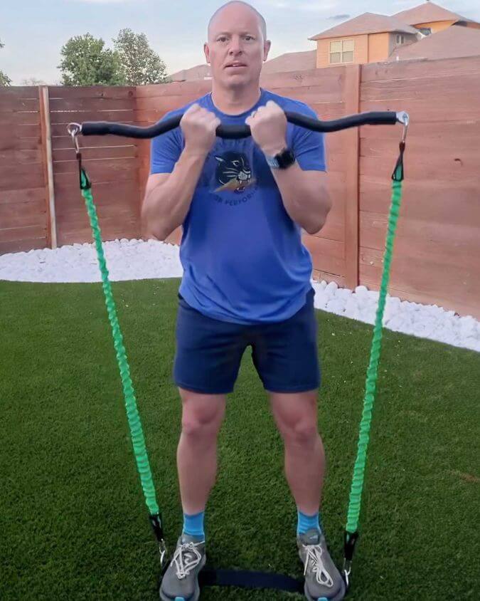 Man performing an exercise with a xbar fitness exercise bar using 30 pound green resistance bands and extension attachments outside.