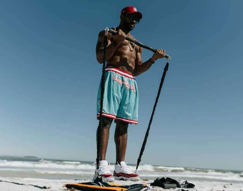 Man standing on a beach performing a bicep curl using xbar bar and resistance system 2.0 bands, wearing turquoise shorts with red and white accents.