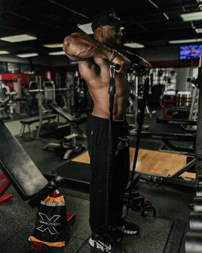 Man performing an upright row exercise with a xbar fitness exercise bar using looped 70 pound black resistance bands and extension attachments creating massive tension.