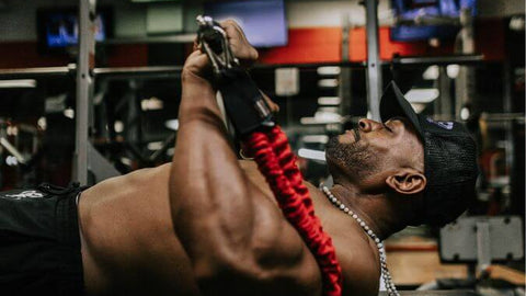 Person using red 50 pound resistance bands, looped 100 pounds, on a gym bench with gym equipment in the background.