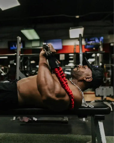 Person using red 50 pound resistance bands, looped 100 pounds, on a gym bench with gym equipment in the background.