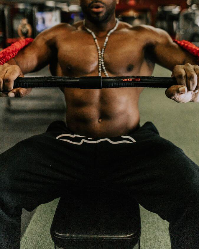 Man performing a chest press with a xbar fitness exercise bar using 50 pound looped red resistance bands creating 100 pounds of tension.