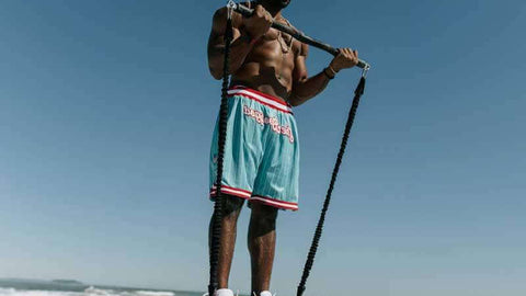 Man standing on a beach performing a bicep curl using xbar bar and resistance system 2.0 bands, wearing turquoise shorts with red and white accents.