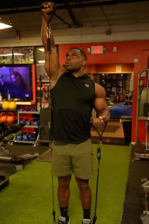 Man exercising with heavy duty handles with resistance bands in a gym setting performing an overhead press.