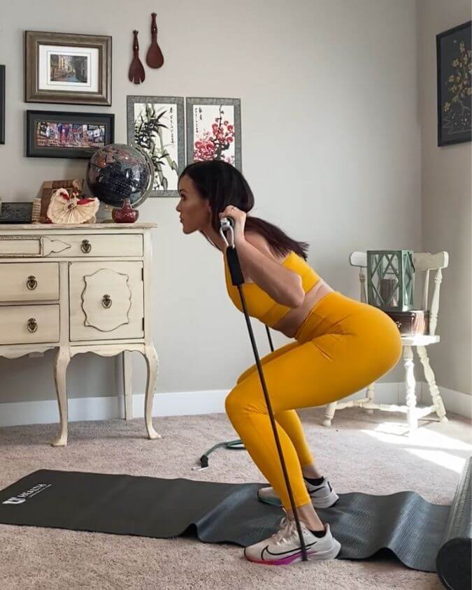 Woman using xbar exercise bar with heavy resistance black band with 70lbs of tension in her living room or spare bedroom performing a squat to show you can workout and exercise anywhere.