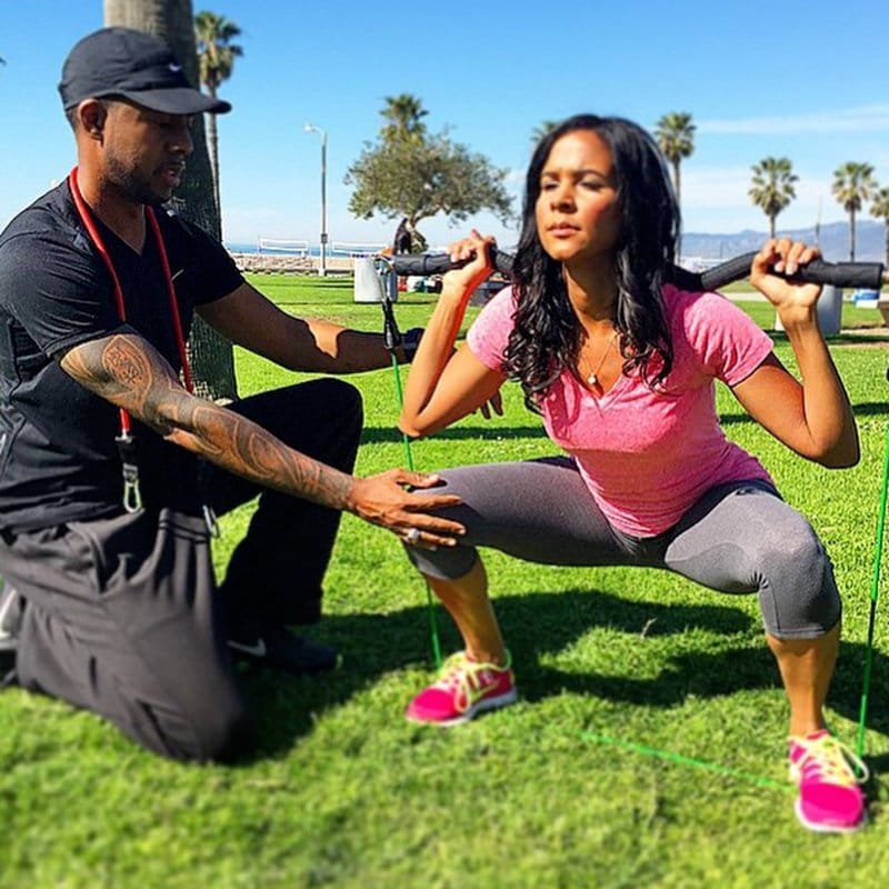 Personal trainer assists woman with resistance band squats in a sunny park setting with palm trees.