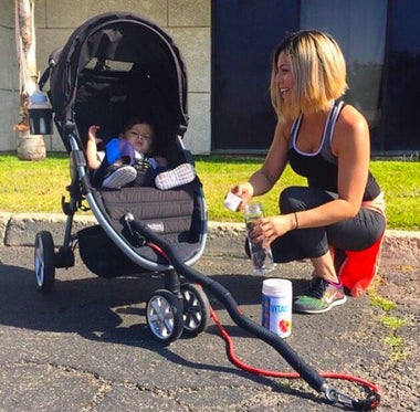 A mom laughs while seated next to her baby in a stroller, holding a water bottle and a supplement can.